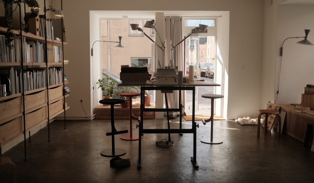 A desk with a few books and two stools is in the center of a room with a large window.